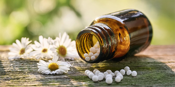 Braunes Glasfläschchen liegt auf einer Holzoberfläche, weiße Kügelchen sind herausgefallen, daneben mehrere Kamillenblüten.